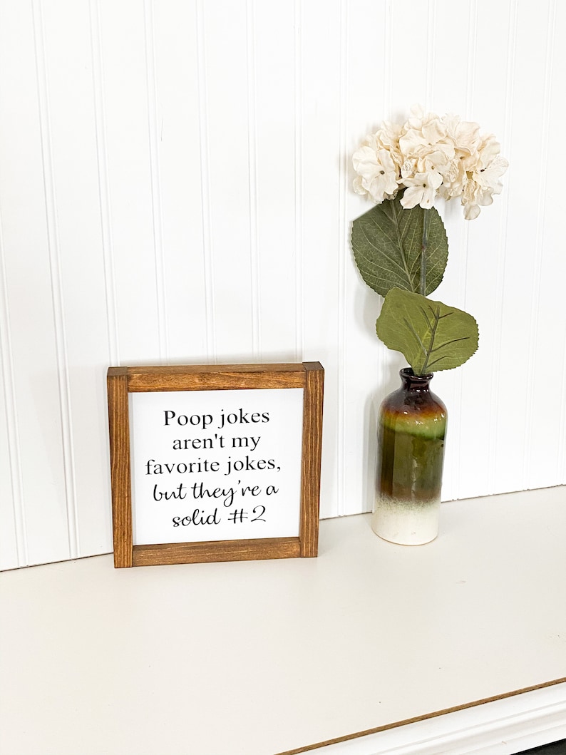 May include: A wooden sign with the text "Poop jokes aren't my favorite jokes, but they're a solid #2". The sign is sitting on a white surface with a white flower in a brown and green vase.