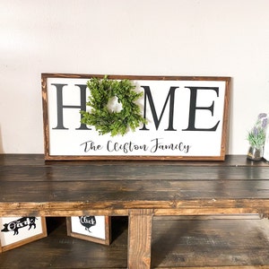 May include: A rectangular wooden sign with the word "HOME" in large black letters on a white background. A small green wreath is centered on the "O". Below, the sign reads "The Clifton Family" in script. The sign has a dark brown frame.