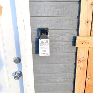 May include: A security camera and a sign that reads "PLEASE REMOVE YOUR SHOES AT THE DOOR! THANK YOU!" are mounted on a gray wooden wall. A white door and a wooden fence are also visible.