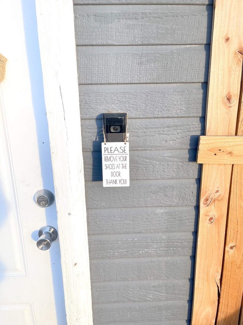 May include: A security camera and a sign that reads "PLEASE REMOVE YOUR SHOES AT THE DOOR! THANK YOU!" are mounted on a gray wooden wall. A white door and a wooden fence are also visible.