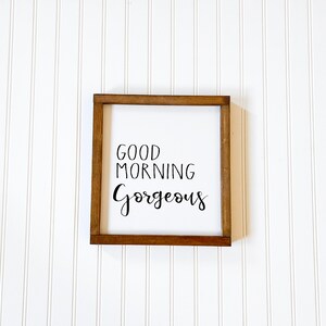 May include: A wooden sign with a white background and brown frame. The sign says "Good Morning Gorgeous" in black cursive lettering.
