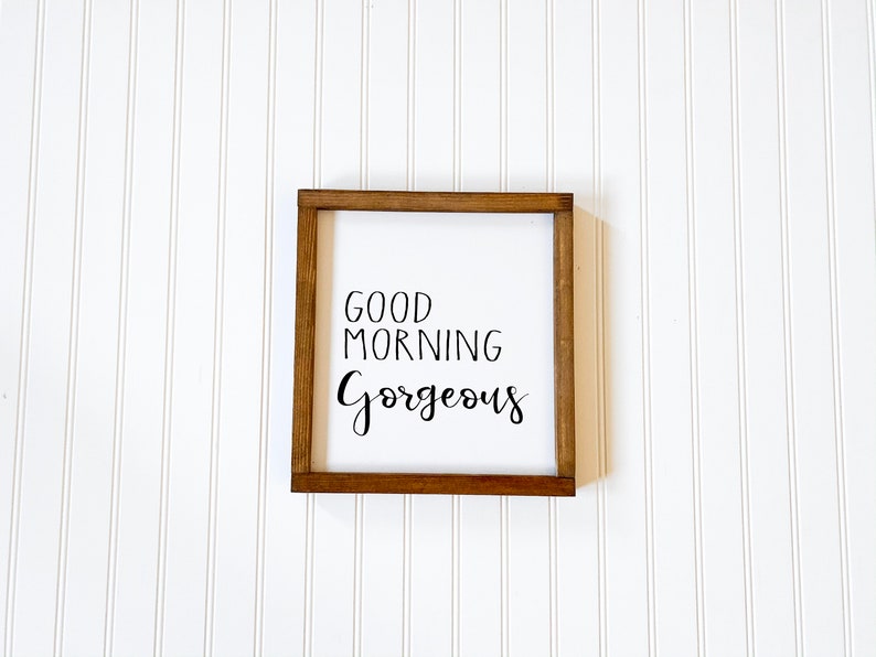 May include: A wooden sign with a white background and brown frame. The sign says "Good Morning Gorgeous" in black cursive lettering.