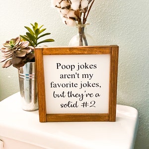 May include: A wooden framed sign with the text "Poop jokes aren't my favorite jokes, but they're a solid #2". The sign is sitting on a white toilet tank. There are plants and a vase with cotton in the background.