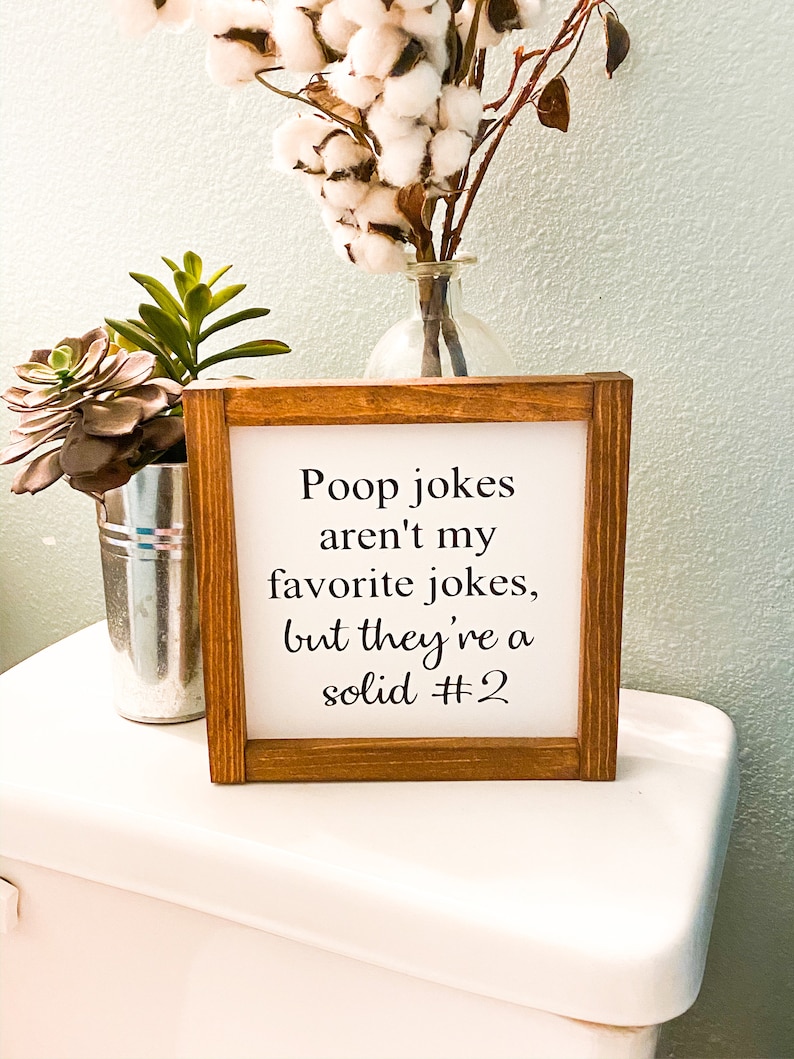 May include: A wooden framed sign with the text "Poop jokes aren't my favorite jokes, but they're a solid #2". The sign is sitting on a white toilet tank. There are plants and a vase with cotton in the background.