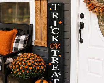 May include: A tall, rectangular black wooden sign featuring the vertical white text TRICK OR TREAT. The design includes small orange pumpkin icons, white ghost figures, and candy corn graphics interspersed between the letters on the sign.
