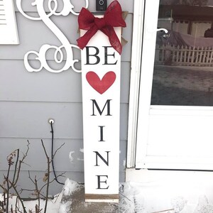May include: White wooden sign with the words "BE MINE" in black lettering and a red heart. The sign is decorated with a red burlap bow and has a rustic twine hanger.