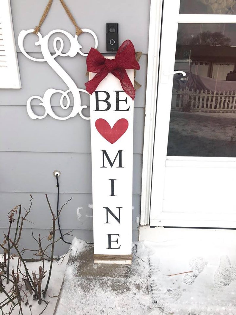 May include: White wooden sign with the words "BE MINE" in black lettering and a red heart. The sign is decorated with a red burlap bow and has a rustic twine hanger.