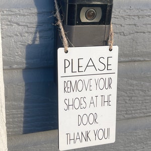 May include: A black video doorbell with a camera lens is mounted on a gray wall. A white rectangular sign with black text that reads "PLEASE REMOVE YOUR SHOES AT THE DOOR. THANK YOU!" is hanging from the doorbell.