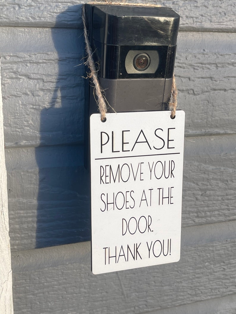 May include: A black video doorbell with a camera lens is mounted on a gray wall. A white rectangular sign with black text that reads "PLEASE REMOVE YOUR SHOES AT THE DOOR. THANK YOU!" is hanging from the doorbell.