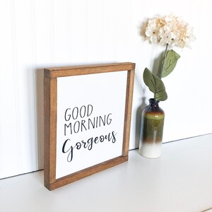 May include: A wooden framed sign with the text "GOOD MORNING GORGEOUS" in black script font. The sign is sitting on a white surface next to a vase with white flowers.