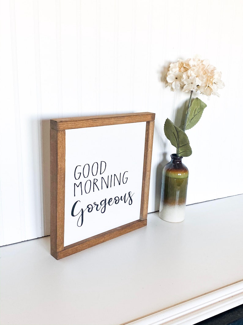 May include: A wooden framed sign with the text "GOOD MORNING GORGEOUS" in black script font. The sign is sitting on a white surface next to a vase with white flowers.
