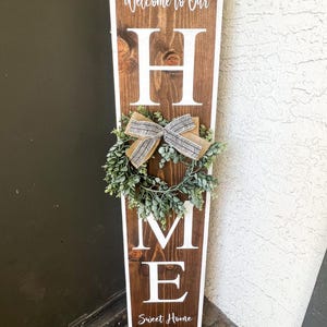 May include: A wooden sign with the word "HOME" in white letters. The sign is decorated with a small wreath and a bow. The sign also says "Welcome to Our" and "Sweet Home".