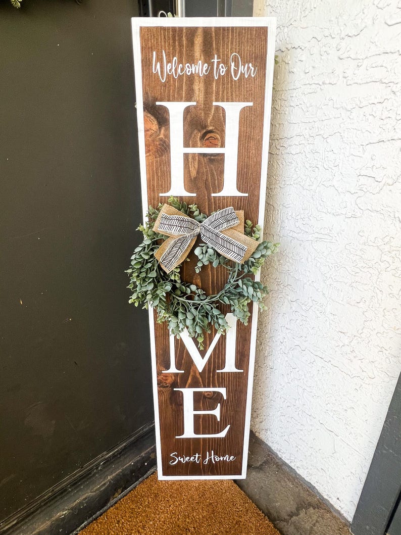 May include: A wooden sign with the word "HOME" in white letters. The sign is decorated with a small wreath and a bow. The sign also says "Welcome to Our" and "Sweet Home".