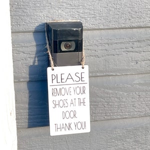 May include: A black video doorbell with a camera lens, mounted on a gray wooden wall. A small white sign is attached with twine, displaying the text: "PLEASE REMOVE YOUR SHOES AT THE DOOR. THANK YOU!"