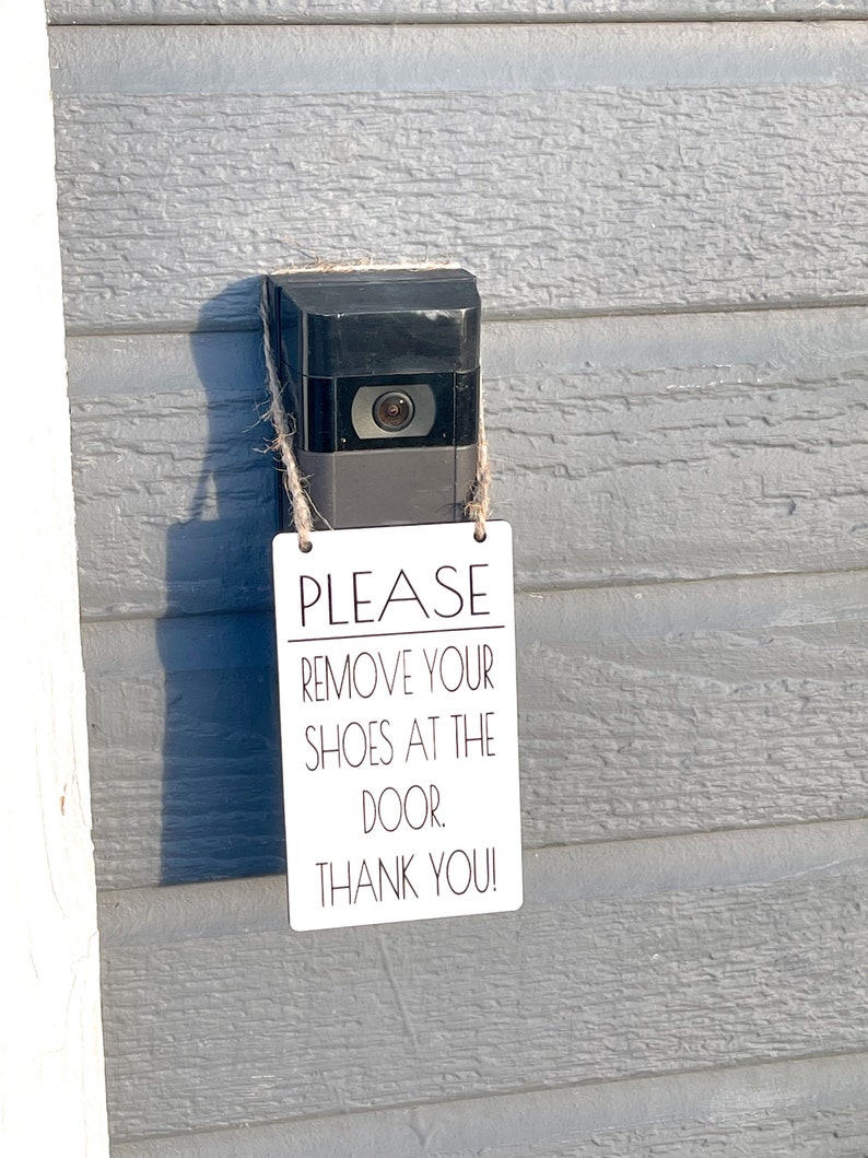 May include: A black video doorbell with a camera lens, mounted on a gray wooden wall. A small white sign is attached with twine, displaying the text: "PLEASE REMOVE YOUR SHOES AT THE DOOR. THANK YOU!"