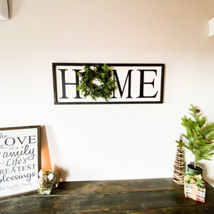 May include: A wooden sign with the word "HOME" in large black letters, with a green wreath in the center. A framed sign with the text "The LOVE of a Family is one of Life's Greatest Blessings" is on a dark wooden table, along with a small Christmas tree.