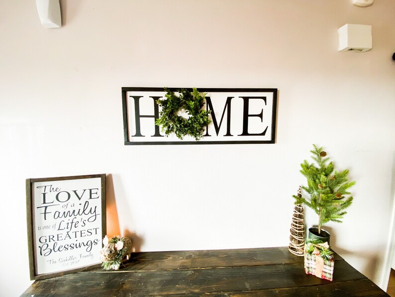 May include: A wooden sign with the word "HOME" in large black letters, with a green wreath in the center. A framed sign with the text "The LOVE of a Family is one of Life's Greatest Blessings" is on a dark wooden table, along with a small Christmas tree.