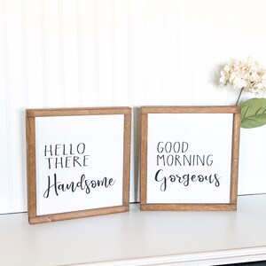May include: Two wooden framed signs with white backgrounds and black calligraphy. The left sign says "Hello There Handsome" and the right sign says "Good Morning Gorgeous".