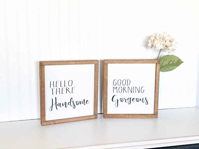 May include: Two wooden framed signs with white backgrounds and black calligraphy. The left sign says "Hello There Handsome" and the right sign says "Good Morning Gorgeous".
