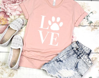 May include: A pink t-shirt with a white paw print and the word "LOVE" printed on the front. The shirt is laying on a white wooden surface with a pair of white shoes and denim shorts.