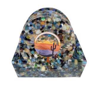 May include: Set of four round coasters with a colorful pebble design, resting in a matching tray. Each coaster features a desert landscape illustration with a cactus. The coasters and tray are made of resin.