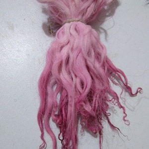 May include: A bundle of pink and white wool fibers, tied together with a string. The fibers are long and curly, and they are arranged in a loose, flowing shape.