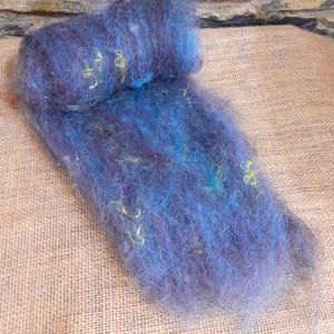 Könnte beinhalten: Nahaufnahme eines Bündels aus blauer und violetter Wollroving mit gelben Fasersträngen. Die Wolle ist weich und flauschig, mit leicht irisierendem Glanz. Das Roving wird auf einer beigefarbenen Jutefläche präsentiert.