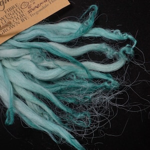 May include: A bundle of teal and white alpaca fiber, labeled "gaver Fibre" with details about the fiber's origin, weight, and length. The label also includes the name "SPEARMINT" and the grower, "Tregondale Flock".