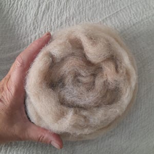Op de afbeelding: Een bal van lichtbeige wolvlies, strak gewikkeld en getextureerd. De wol is zacht en pluizig, ideaal voor verschillende vezelambachten zoals vilten en spinnen. De neutrale kleur maakt het veelzijdig.