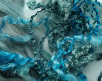 May include: Close-up of a pile of dyed wool fibers in shades of blue and teal. The fibers vary in texture, with some being straight and others curly. The colors range from light blue to deep teal, creating a visually interesting display.