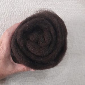 Op de afbeelding: Een donkerbruine wolvlies, strak opgerold in een spiraalvorm. De wol wordt in een hand gehouden en ziet er zacht en pluizig uit. De achtergrond is een lichte, getextureerde stof.