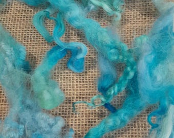 May include: Close-up of vibrant, hand-dyed wool roving in shades of turquoise and teal. The soft, fluffy fibers are arranged in swirling patterns, creating a textured and artistic display. Ideal for fiber art projects.