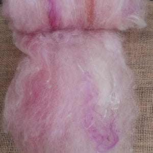 Op de afbeelding: Een massa zachte, lichtroze vezels, mogelijk wol of een vergelijkbaar textiel, met paarse en oranje strepen. Het materiaal lijkt in een ruwe staat te zijn, geschikt voor handwerk zoals vilten of spinnen. De textuur is zacht en pluizig.