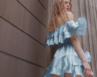 May include: A woman wearing a light blue off-the-shoulder dress with multiple ruffles. The dress is short and has a flowy, feminine silhouette.