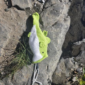 May include: A handmade crochet fish toy featuring a bright lime green body with a white underbelly, small side fins, and a dark circular eye. The fish is attached to a white cord loop at the tail end, showcasing a textured yarn construction.