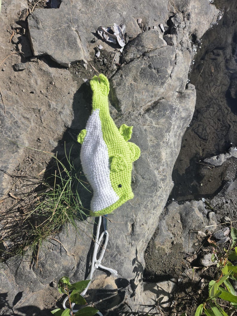 May include: A handmade crochet fish toy featuring a bright lime green body with a white underbelly, small side fins, and a dark circular eye. The fish is attached to a white cord loop at the tail end, showcasing a textured yarn construction.