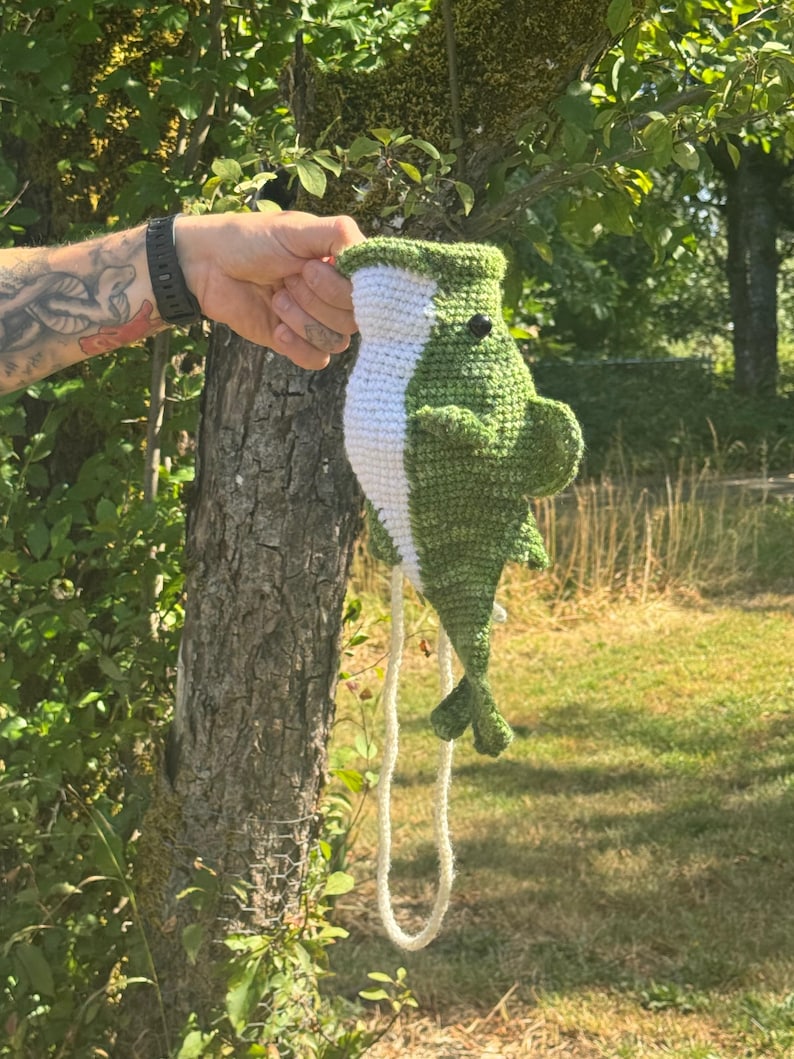 May include: A crocheted fish toy featuring a dark green body with a white underbelly, a small black eye, side fins, and a tail. The fish has a long, thin white cord attached to its lower section, allowing it to hang vertically from a top opening.