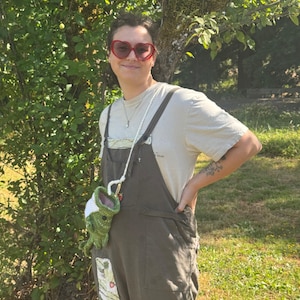 May include: A pair of red heart-shaped sunglasses, a beige short-sleeved t-shirt, and dark olive green overalls with a patch pocket. A small, green, crocheted frog-shaped pouch with a white top hangs from a long, thin, white cord strap.