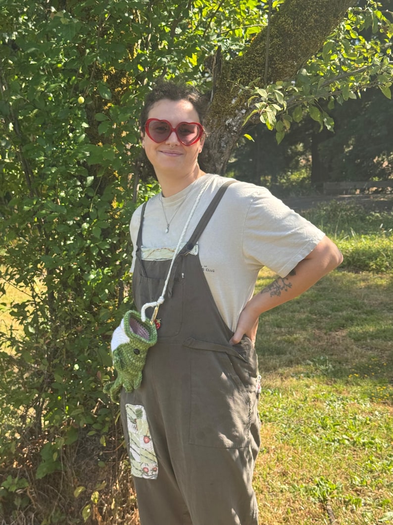 May include: A pair of red heart-shaped sunglasses, a beige short-sleeved t-shirt, and dark olive green overalls with a patch pocket. A small, green, crocheted frog-shaped pouch with a white top hangs from a long, thin, white cord strap.