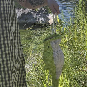 May include: A crochet bag shaped like a fish with a light green body, a white underbelly, and a small black eye. The bag features a top opening and is attached to a white cord with a metal carabiner clip for carrying or hanging purposes.