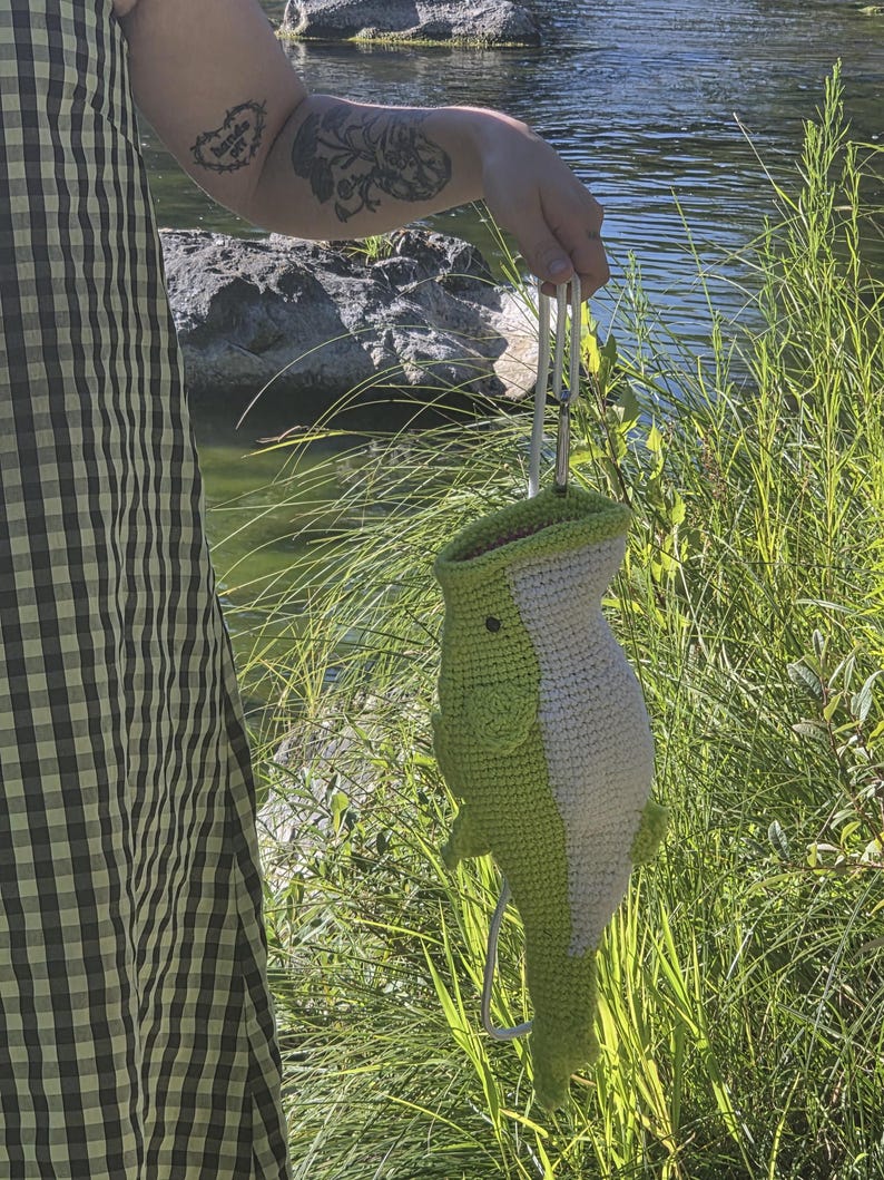 May include: A crochet bag shaped like a fish with a light green body, a white underbelly, and a small black eye. The bag features a top opening and is attached to a white cord with a metal carabiner clip for carrying or hanging purposes.