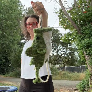 May include: A handmade crochet plush toy shaped like a shark, featuring a dark green back, a white underbelly, a small black eye, and side fins. The shark is suspended by a white yarn loop attached to the top, showcasing its vertical orientation.