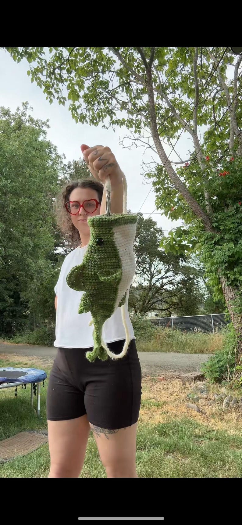 May include: A handmade crochet plush toy shaped like a shark, featuring a dark green back, a white underbelly, a small black eye, and side fins. The shark is suspended by a white yarn loop attached to the top, showcasing its vertical orientation.