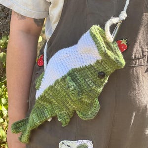 May include: A crochet fish-shaped bag featuring a green body with a white belly, a black eye, and fins. The bag has a white yarn strap attached to the top opening, which is held open by a metal crochet hook. The fish is textured with yarn stitches.