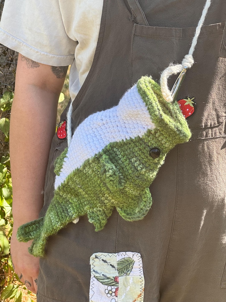 May include: A crochet fish-shaped bag featuring a green body with a white belly, a black eye, and fins. The bag has a white yarn strap attached to the top opening, which is held open by a metal crochet hook. The fish is textured with yarn stitches.