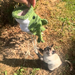 May include: A green and white crocheted fish toy with a wide open mouth, black plastic eyes, and two long off-white strings hanging from the bottom, featuring a textured yarn construction and a distinct fin shape on the side of the body.