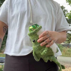 May include: A green and white crocheted drink holder shaped like a dinosaur with a long tail and a white underbelly. The holder features a black eye and is designed to carry a standard aluminum beverage can, suspended by a white cord strap.