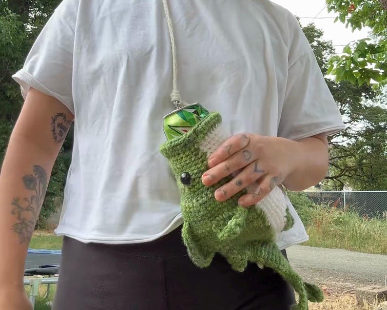 May include: A green and white crocheted drink holder shaped like a dinosaur with a long tail and a white underbelly. The holder features a black eye and is designed to carry a standard aluminum beverage can, suspended by a white cord strap.