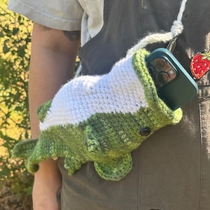 May include: Crocheted green and white phone case shaped like a fish, with a black button eye. The case has a white strap and a phone inside. A small strawberry patch is visible on the strap.