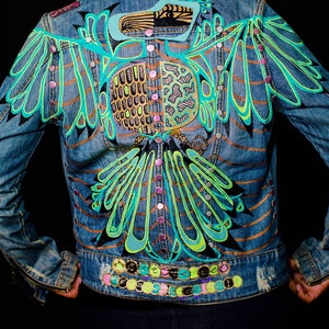 May include: A denim jacket with a hand-painted design of a stylized bird with green, blue, and gold accents. The jacket has a row of silver buttons and a row of colorful embellishments at the bottom.